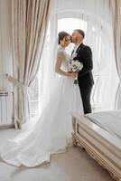 a joyful couple of young brides hug, kiss in a hotel room. The bride in a wedding dress hugs the cheerful groom in classic formal clothes by the shoulder. The first meeting of the bride and groom photo