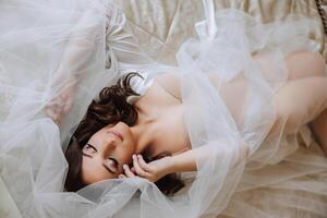 un hermosa morena novia con un tiara en su pelo es preparando para el Boda en un hermosa túnica en tocador estilo. de cerca Boda retrato de el novia acostado en el cama, foto desde arriba.