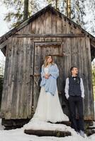 Groom and bride on the background of a wooden forest house. The bride is wrapped in a blue blanket. Winter wedding. photo