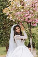 Curly brunette bride in a lush veil and long-sleeved dress poses near cherry blossoms. Spring wedding photo