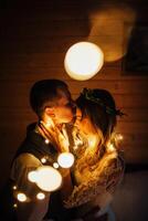 a happy bride and groom embrace in a dark room illuminated by defocused lights of a garland photo
