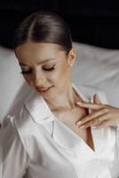 un sensual y seguro de sí mismo novia es sentado en un cama en blanco pijama en un hotel habitación. moderno Corte de pelo y constituir. Boda concepto. Mañana de el novia. preparación para el Boda ceremonia foto