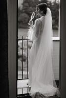 A beautiful bride with a long veil on her hair, wearing an elegant white peignoir, standing on a balcony overlooking the city, drinking a drink during the wedding morning. black and white photo