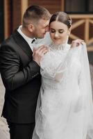 Portrait. A bride in an elegant long dress and veil and a groom in a suit pose while standing near trees. Wedding in nature photo