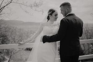 Black and white portrait. Bride in elegant long dress and groom in black suit hugging and posing near wooden railing. Wedding in nature photo