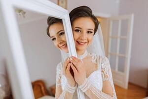 A bride in an elegant dress with long sleeves poses by a mirror in her room. photo