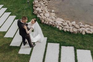 Portrait. The bride in an elegant long dress and the groom are hugging and posing in the yard, standing on a stone path. Wedding in nature photo