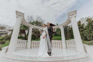 A bride in a long dress and a groom in a black suit are standing near white Roman-style columns. Beautiful hair and makeup. An exquisite wedding photo