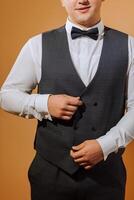 a young smiling man in a white shirt wears a bow tie and a black waistcoat. A groom with a short hairstyle on an orange background puts on his black suit and prepares for the wedding photo