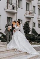 un novia en un blanco vestir con un tren y un novio en un traje actitud en el pasos de un edificio. Boda foto sesión en naturaleza