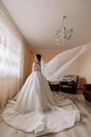 The bride in a long, voluminous wedding dress poses in her room. The veil is thrown into the air. Long train. Portrait of the bride. Open shoulders and neckline. photo