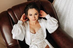 A bride in a lace bodysuit and silk robe sits and poses in a leather chair in her room. Morning of the bride photo