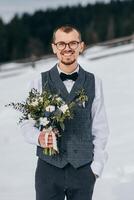 Wedding portrait of the groom. The groom stands against the background of the winter forest. A man in a vest and white shirt, glasses and a bow tie. Winter wedding photo