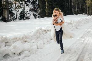 el novia y novio son corriendo a lo largo un Nevado la carretera en contra el antecedentes de un pino bosque y hermosa contraste luz de sol. lado vista. invierno boda. sitio para logo. foto