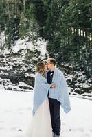 Groom and bride against the background of a pine forest and mountains. The bride and groom are wrapped in a blue blanket. Winter wedding. photo