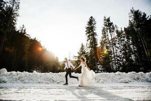 The bride and groom are running along a snowy road against the background of a pine forest and beautiful contrast sunlight. Side view. Winter wedding. Place for logo. photo