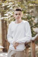 Close-up vertical portrait of a teenager in a white sweater and brown pants. Happy smiling teenager in summer park in sunlight. A beautiful child is looking at the camera in the clearing. photo
