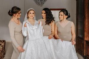 The bride in a white robe and her friends in cream dresses pose near the bride's dress. Morning of the bride photo