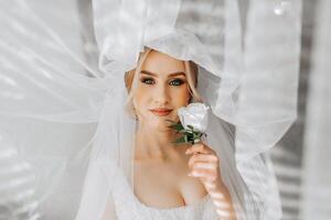 el novia en un blanco vestido, cubierto con un velo, poses en bueno Encendiendo en su habitación. Mañana de el novia. un hermosa tiara foto