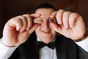 the groom holds the wedding rings with his fingers at the level of his eyes and looks into the camera through the rings. Funny photo. Selective focus. photo