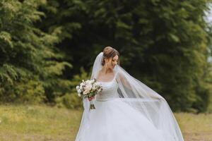 The bride in a lush dress and veil, holding a bouquet of white flowers and greenery, poses with the veil thrown in the air, against the background of green trees. Spring wedding photo