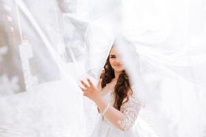 Curly brunette bride poses wrapped in a veil against the background of mountains and wooden houses. Magnificent dress with long sleeves, open bust. Summer wedding photo