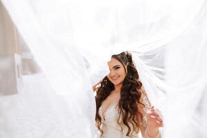 Curly brunette bride poses wrapped in a veil against the background of mountains and wooden houses. Magnificent dress with long sleeves, open bust. Summer wedding photo