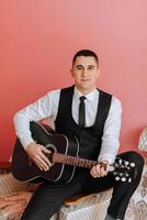 The groom sings songs to the guitar in the morning in his room. Groom in a black suit. Happy man. Preparation for the wedding. photo