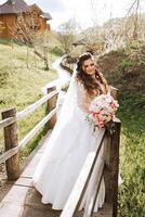 Curly brunette bride poses with a bouquet, near a wooden railing. Magnificent dress with long sleeves, open bust. Spring wedding photo