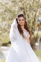 Curly brunette bride poses wrapped in a veil against the background of mountains and wooden houses. Magnificent dress with long sleeves, open bust. Summer wedding photo