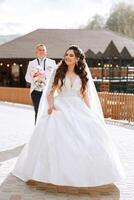 A beautiful young bride, in a summer park, walks ahead of her groom. Beautiful wedding white dress. Walks in the park. A happy and loving couple. photo