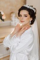 Morning of the bride before the wedding. A beautiful young woman with a veil and a beautiful hairstyle in a white robe in a room with a wonderful interior. Natural beauty and professional makeup. photo