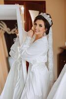 portrait of a luxurious bride in boudoir style near a mirror with reflection. Preparing the bride for the wedding in the morning in a white robe. photo