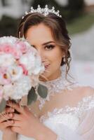 brunette bride in off-the-shoulder lace white dress and tiara posing with a bouquet of white and pink flowers. Beautiful hair and makeup. Spring wedding photo