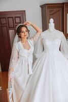 A happy bride is preparing for her luxurious wedding in a hotel room, with a wedding dress on a mannequin nearby. Portrait of a woman with fashionable hair, makeup and a smile in a dressing gown. photo