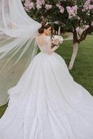 brunette bride in a lacy white dress with a long train, posing with a bouquet of white and pink flowers, against the background of trees. The veil is in the air. Beautiful hair and makeup. photo