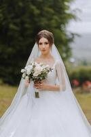 The bride in a lush dress and veil, holding a bouquet of white flowers and greenery, posing against the background of green trees.. Spring wedding photo