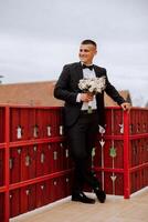 The groom in a black suit holds a bouquet, poses leaning on a red railing. Wedding portrait. photo