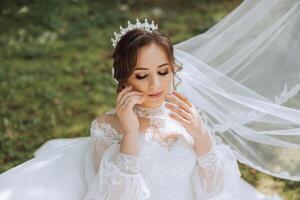 The bride in a lush dress with long sleeves poses with her veil in the air, posing while sitting on the grass, against a green background. Beautiful hairstyle, royal tiara. Spring wedding photo