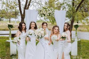 Group portrait of the bride and bridesmaids. Bride in a wedding dress and bridesmaids in white dresses and holding stylish bouquets on the wedding day against the background of the lake. photo