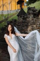 A curly-haired brunette bride in a lace dress with an open bust poses against the background of a stone building, throwing her veil in the air. Spring wedding photo