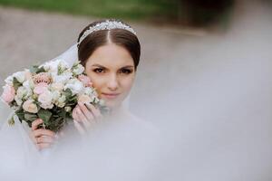 brunette bride in a lacy white dress with a long train, posing with a bouquet of white and pink flowers, against the background of trees. The veil is in the air. Beautiful hair and makeup. photo