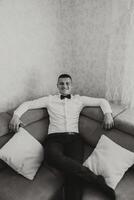 elegante retrato de el novio preparando para el Boda ceremonia. del novio Mañana. el novio es sentado en el sofá. el confidente Mira de un maduro hombre. negro y blanco foto. foto
