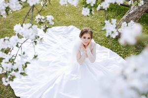 Against the background of a blooming tree, a red-haired bride poses, sitting on green grass. Magnificent dress with long sleeves, open bust. Spring wedding photo