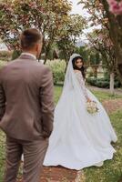 A beautiful young bride, in a summer park, walks ahead of her groom. Beautiful wedding white dress. Walks in the park. A happy and loving couple. photo