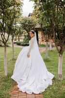 brunette bride in a lacy white dress with a long train, posing with a bouquet of white and pink flowers, against the background of trees. The veil is in the air. Beautiful hair and makeup. photo