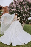 brunette bride in a lacy white dress with a long train, posing with a bouquet of white and pink flowers, against the background of trees. The veil is in the air. Beautiful hair and makeup. photo