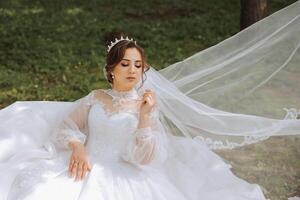 The bride in a lush dress with long sleeves poses with her veil in the air, posing while sitting on the grass, against a green background. Beautiful hairstyle, royal tiara. Spring wedding photo