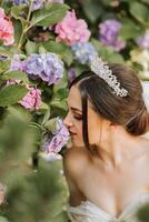 young beautiful bride in wedding dress with open shoulders and crown on her head sitting near hydrangea flowers, fashion photo taken under harsh sunlight