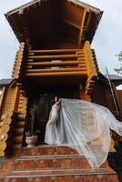 A curly-haired brunette bride in a lace dress with an open bust poses against the background of a wooden house, throwing her veil in the air. Spring wedding photo